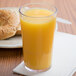 A Cambro clear plastic tumbler filled with orange juice next to a bagel on a table.