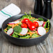 A black 10 Strawberry Street Wazee Matte stoneware bowl filled with salad with tomatoes, radishes, and spinach.