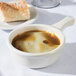 A Tuxton eggshell white French casserole bowl of brown soup with a piece of bread on a white table.
