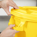 A hand opening a yellow Rubbermaid BRUTE trash can lid.