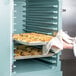 A person holding trays of food in a Cambro double compartment tray and sheet pan carrier over an open oven.
