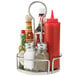 A Tablecraft stainless steel condiment caddy on a metal stand holding condiments in glass jars and bottles.