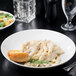 A Libbey porcelain pasta bowl filled with pasta on a table with salad and a glass of wine.