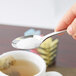 A hand holding a Libbey stainless steel teaspoon full of sugar over a cup of liquid.