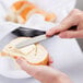 A person using a Reserve by Libbey Baroque stainless steel bread and butter knife to cut a roll of bread.