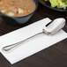 A Libbey extra heavy weight stainless steel serving spoon on a napkin next to a bowl of soup.
