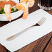 A World Tableware stainless steel cocktail fork on a napkin next to a bowl of shrimp.