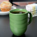 A close-up of a sage green Libbey porcelain mug filled with brown liquid on a white surface with a muffin and cupcake.