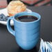 A blue Libbey porcelain mug filled with brown liquid on a black surface with a muffin and blueberries.