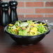 A bowl of salad with vegetables in a black Fineline Super Bowl on a table.