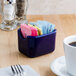 A blue ceramic Tuxton sugar packet holder on a counter with sugar packets in it.