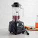 A AvaMix commercial blender on a counter with a glass of juice and a blender filled with fruit.