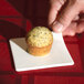 A hand holding a small muffin on a Libbey white porcelain square plate.