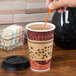 A hand using a Royal Paper Stix To Go orange beverage plug and stirrer to stir coffee in a paper cup with a lid.