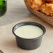 A bowl of white sauce next to fried chicken wings served in a black plastic souffle cup.