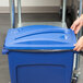 A hand pushing a blue Rubbermaid Slim Jim recycling container lid with a paper slot.