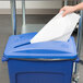 A hand putting a piece of paper into a blue Rubbermaid recycling bin with a paper slot.