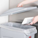 A hand putting paper into a Rubbermaid Slim Jim trash can with a light gray lid.