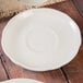 A wood surface with a white Tuxton eggshell scalloped china saucer on a white plate.