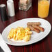 A white oval Elite Global Solutions melamine plate with scrambled eggs and sausages on it.
