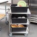 A Lakeside stainless steel utility cart with black containers holding white and green plates.