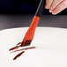 A hand using a Mercer Culinary silicone brush with an orange and black handle to spread brown liquid on a white plate.