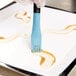 A hand using a blue Mercer Culinary silicone brush to plate brown liquid in a white dish.