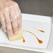 A hand in a plastic glove using a Mercer Culinary silicone wedge to plate brown liquid on a white tray.