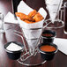 A Clipper Mill stainless steel wire cone basket with 3 ramekins of dipping sauces on a table in a pizza parlor.