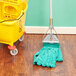 A Rubbermaid green microfiber wet mop in a yellow mop bucket on a wood floor.