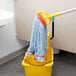 A blue Rubbermaid mop head with a 5" headband on a mop handle in a yellow bucket.