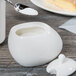 A spoon pours sugar into a white Libbey porcelain sugar pot on a table.