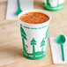 A 32 oz. EcoChoice compostable paper food cup with a tree design filled with soup on a table with a green and white spoon.