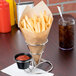 A Clipper Mill stainless steel wire cone basket with ramekin holder holding french fries with ketchup on a table.