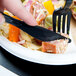 A finger holding a WNA Comet Classicware EcoSense black plastic knife over a plate of food.