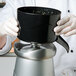 A person in a professional kitchen using a black AvaMix bowl to pour liquid into a blender.