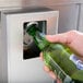 A hand opening a green bottle of beer over a stainless steel horizontal bottle cooler.