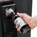 A hand holding a bottle of beer opening a bottle opener above an Avantco horizontal bottle cooler.