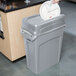 A person putting a white lid on a Rubbermaid grey rectangular trash can.