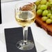 A Fineline clear plastic wine goblet filled with white wine on a black napkin next to grapes.