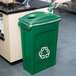 A hand holding a green Rubbermaid recycling bin with a recycle symbol on it.