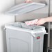 A hand throws a paper into a gray Rubbermaid Slim Jim trash can with a lid.
