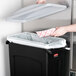 A hand throwing a paper into a Rubbermaid trash can with a light gray lid.