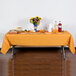 A table with a Creative Converting pumpkin spice orange plastic table cover, food, and flowers on it.