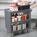 A man holding a Cambro granite gray three shelf service cart with clear plastic containers with red lids on it.