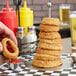 A hand dipping a fried onion ring into a bowl of ketchup on a table with a stack of onion rings in a Clipper Mill stainless steel tower.
