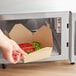 A hand reaching into a microwave with a Kraft paper take-out box filled with a burger, broccoli, and an apple.