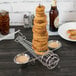 A Clipper Mill stainless steel onion ring tower with ramekin holders filled with onion rings on a table with a bowl of sauce.