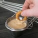 A hand holding a Clipper Mill stainless steel onion ring tower with a fried onion ring over a bowl of sauce.
