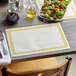 A table set with a Greek Key Gold Choice placemat, a plate of salad, and a glass of wine.